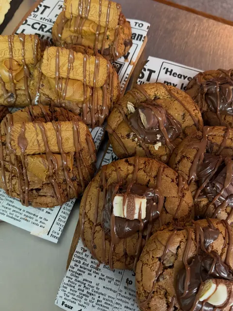 A tray of chunky cookies, some topped with caramel biscuits and others with chocolate pieces, all drizzled with chocolate