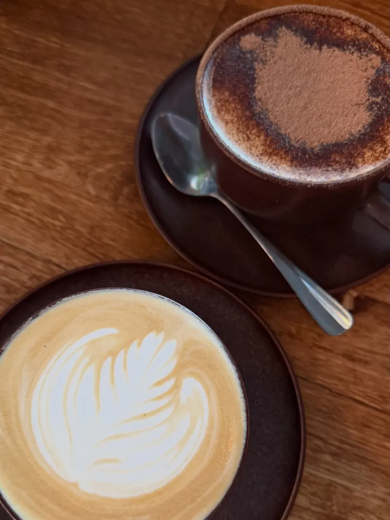 A flat white with leaf latte art beside a hot chocolate on a dark ceramic saucer, on an oak table