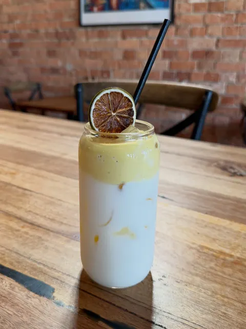 A tall layered iced drink, golden on top and creamy white below, with a dried citrus slice and a black straw, on a timber table by a brick wall