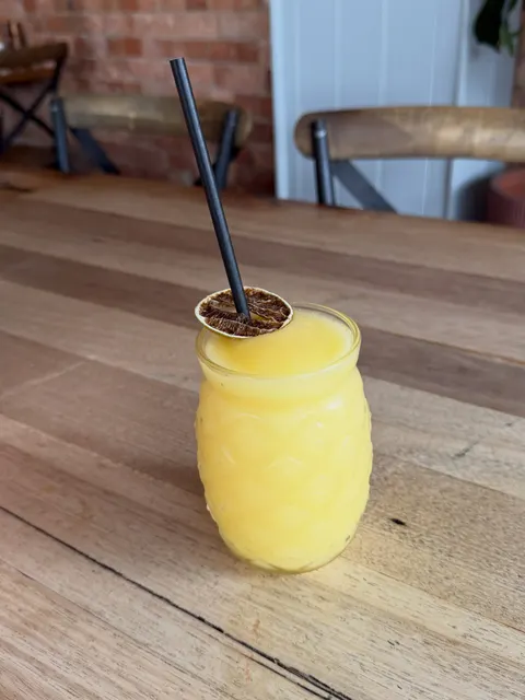 A bright yellow frozen drink in a pineapple-shaped glass with a dried fruit slice and a black straw, on a timber table